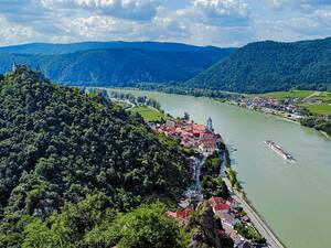 Die Bilderbuchkulisse der Wachau bei Dürnstein mit dem markanten blauen Kirchturm und der Burgruine auf dem Felsen, während ein Kreuzfahrtschiff gemächlich die Donau hinabgleitet.